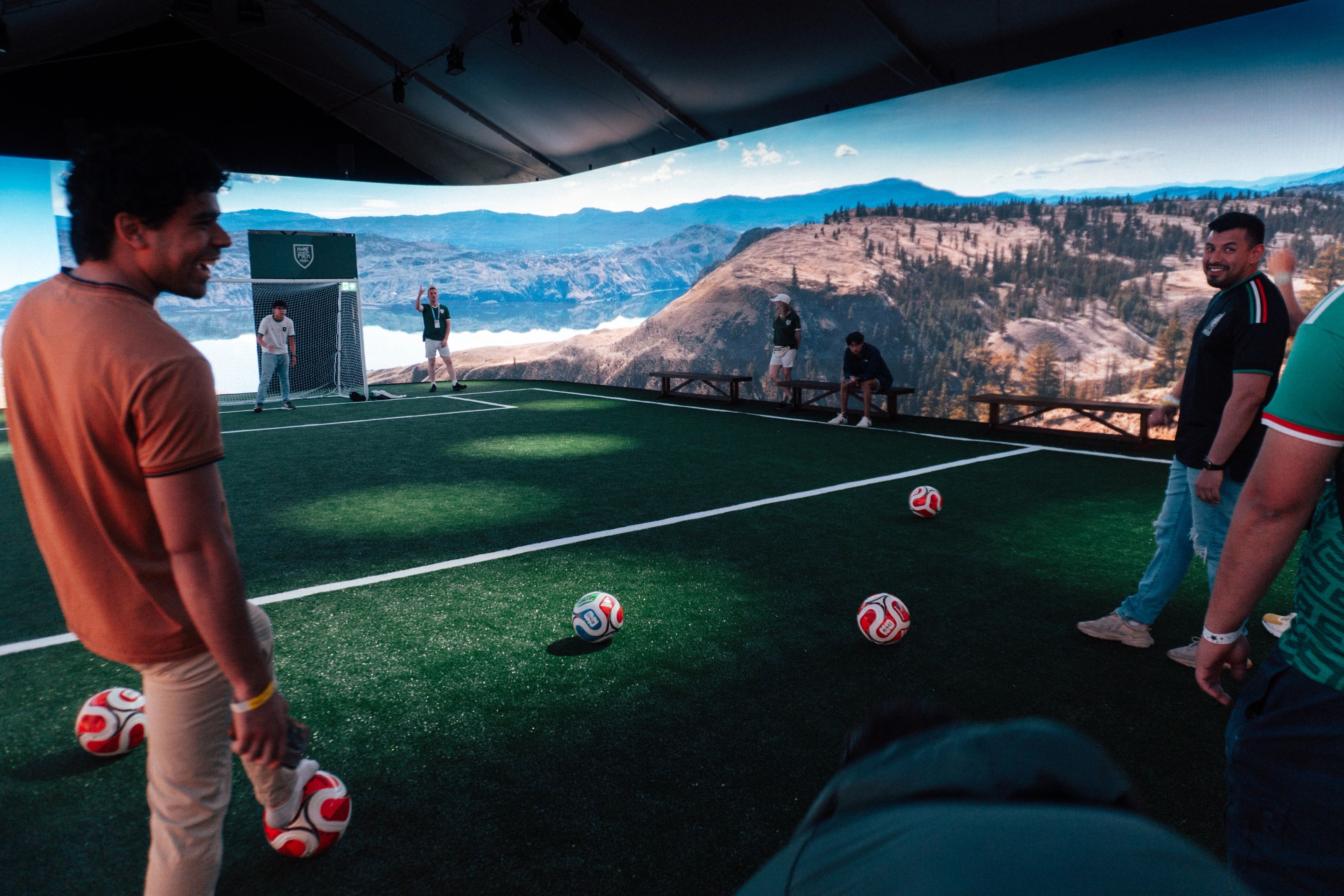 People play soccer on a rooftop turf field overlooking a dramatic mountain valley, with scattered balls and a relaxed, scenic atmosphere.