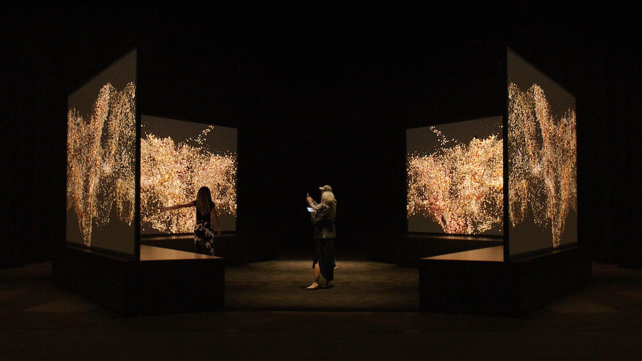 People standing in a dimly lit room surrounded by screens displaying an interactive installation of various shades of Canadian skin tones.