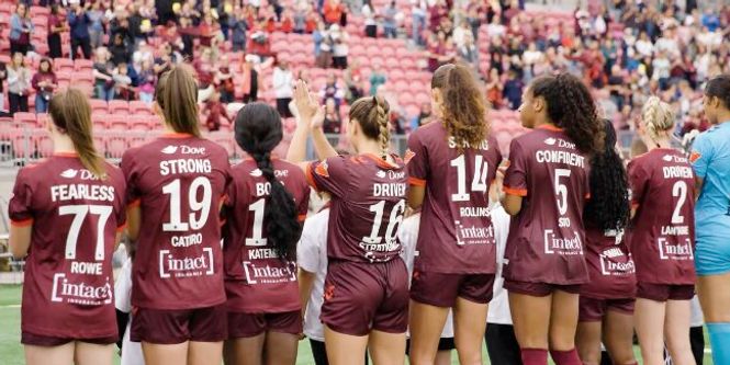 A women’s soccer team in maroon jerseys stands on a field facing cheering fans. The mood is celebratory and supportive, with players and audience engaging enthusiastically.