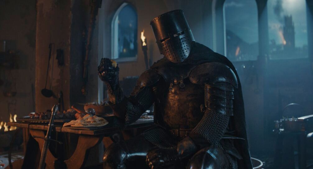 A medieval knight in dark armour sits at a wooden table in a dimly lit room