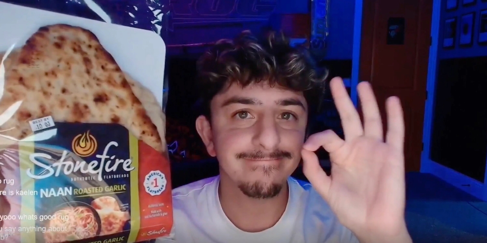 A person with curly hair and a goatee is in a dimly lit room, holding a pack of Stonefire naan bread in one hand and gesturing an "OK" sign with the other, smiling slightly.