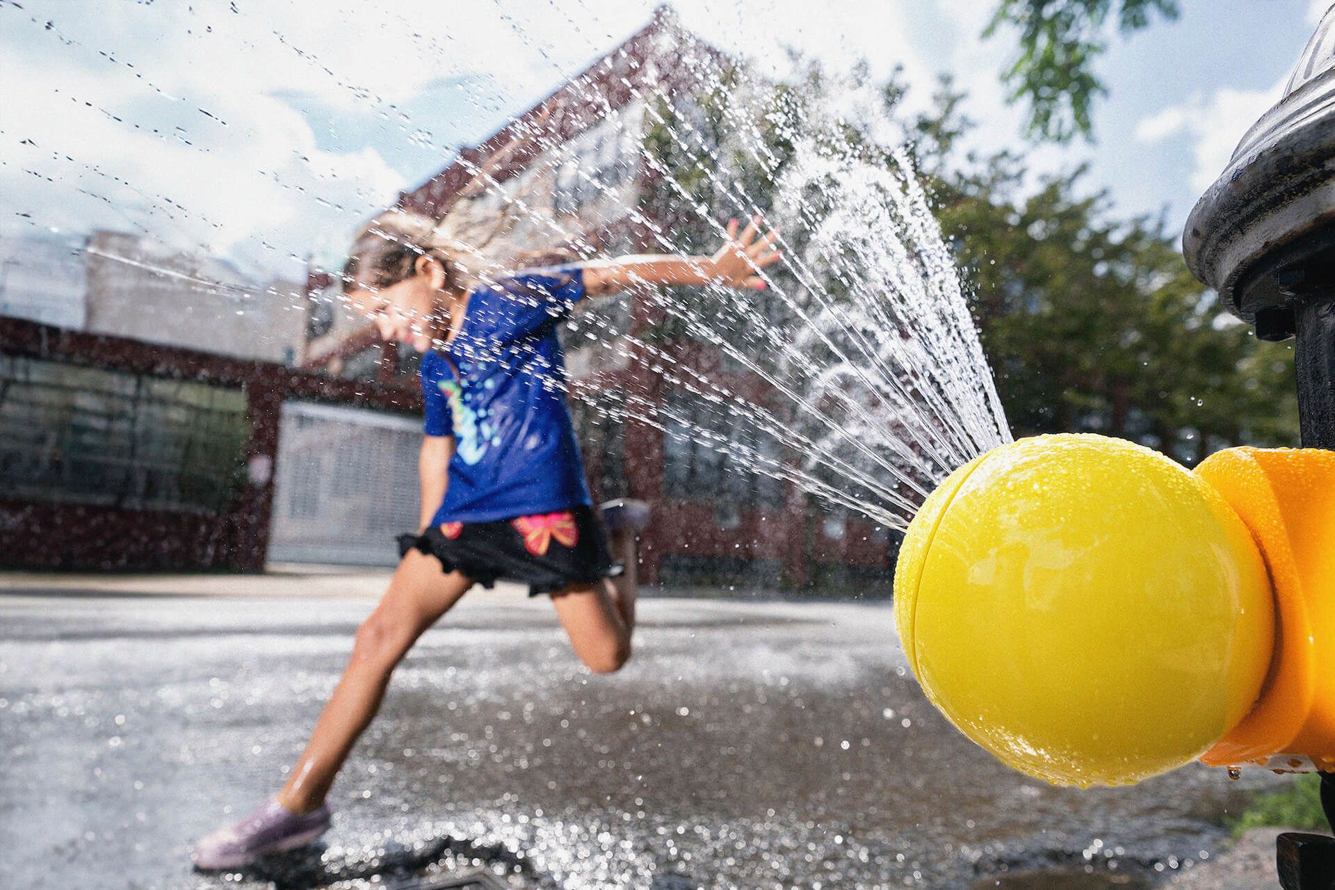A young girl playing in the water coming from a yellow Splash Spot fire hydrant cap on a New York street.