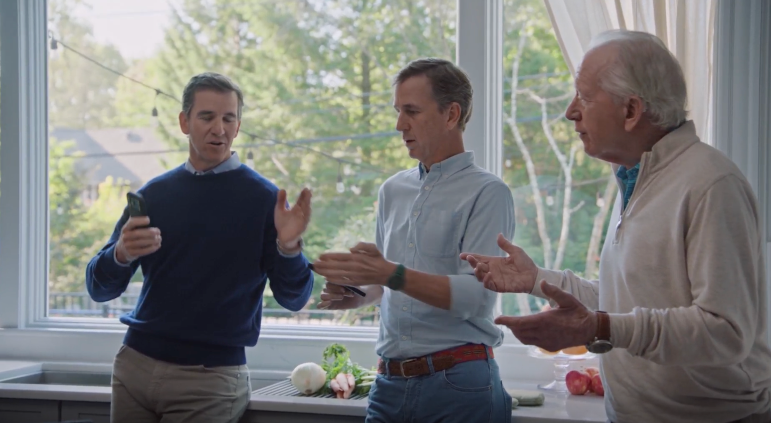 Three men are conversing in a bright kitchen with a large window overlooking greenery.