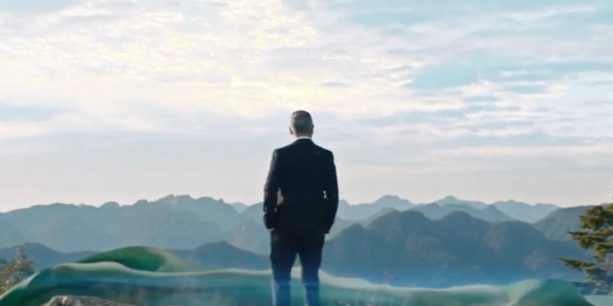 A man in a suit stands atop a rocky mountain, gazing at a vast mountain range under a blue sky with scattered clouds, evoking a sense of reflection.