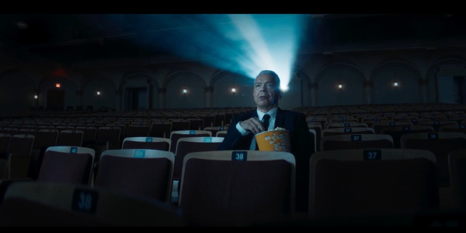 A man sits alone in a dimly lit theater, holding a bucket of popcorn. Projector light beams over his head, creating a nostalgic, cinematic ambiance.