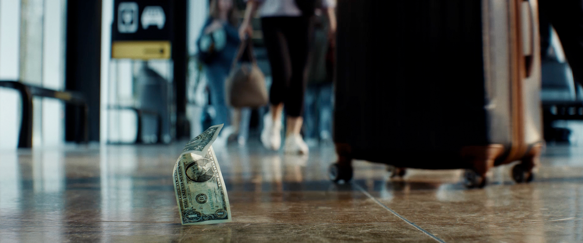 A crumpled US dollar bill stands on a polished airport floor while travelers and rolling luggage pass in the blurred background.