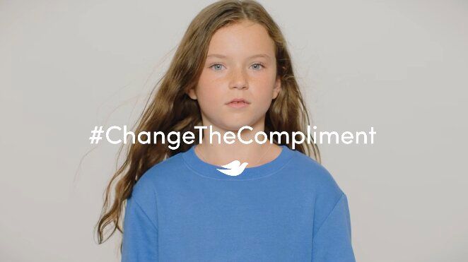 Young girl with long hair in a blue shirt, looking calmly at the camera. The text “#ChangeTheCompliment” is overlaid, promoting positive change.