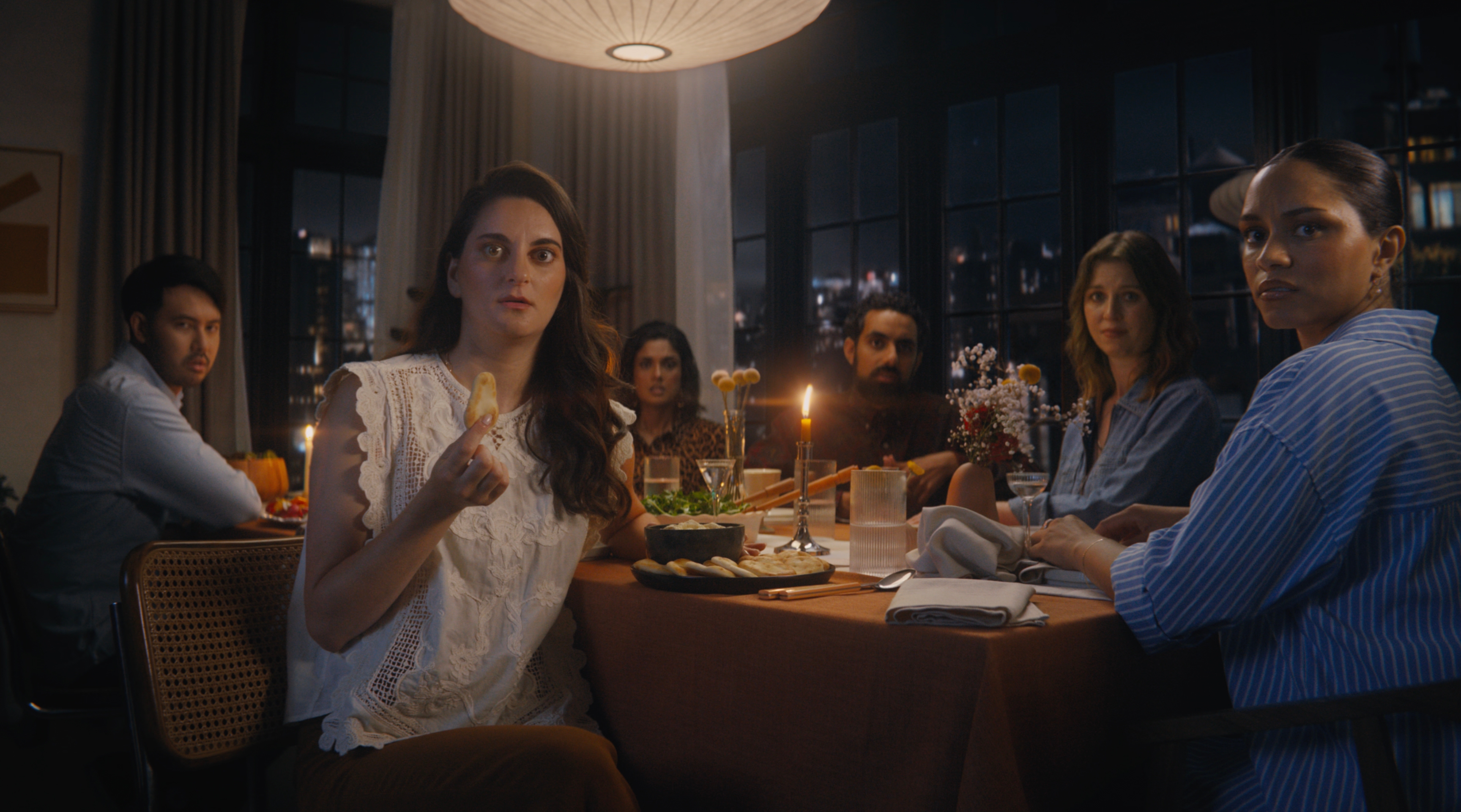 A group of six people sit around a warmly lit dinner table with a candle centerpiece, looking surprised.