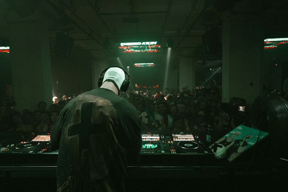 DJ silhouetted from behind at Pioneer booth facing large warehouse crowd