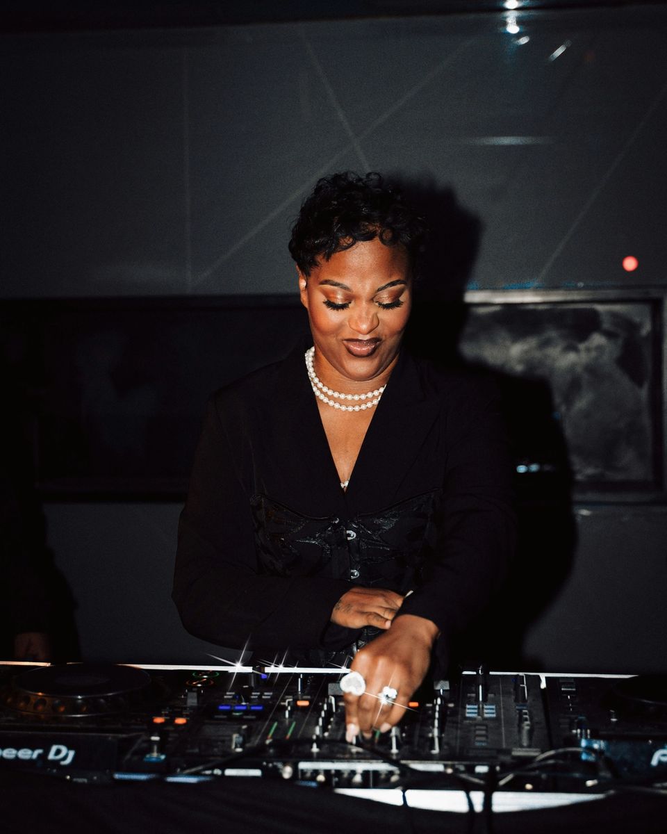 Female DJ performing at a nighttime event, working at a booth in a dark venue with warm lighting