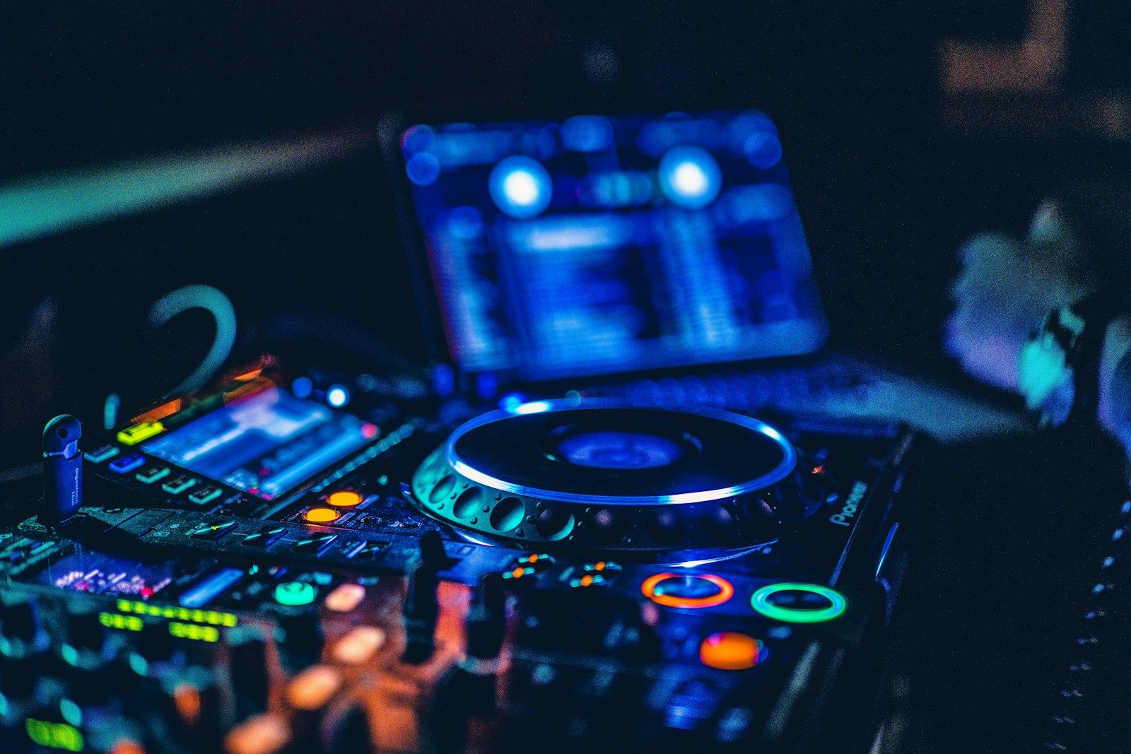 Close-up of a dark DJ booth with a lit CDJ jog wheel, mixer controls and a blurred laptop screen