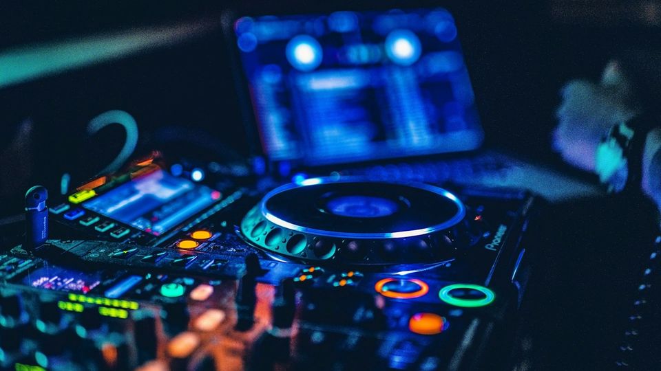 Close-up of a dark DJ booth with a lit CDJ jog wheel, mixer controls and a blurred laptop screen