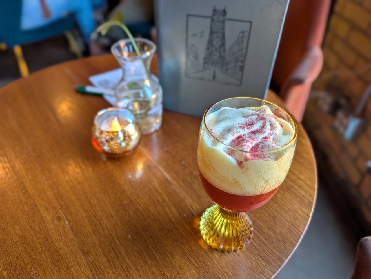 A red cocktail topped with pale foam and pink dust in a yellow-stemmed glass on a table at Vikre