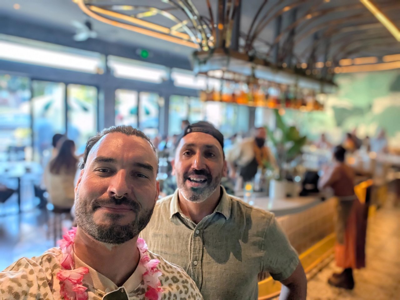 Two friends at Kingfisher, one wearing a pink flower lei, with the curved bar and botanical mural behind them