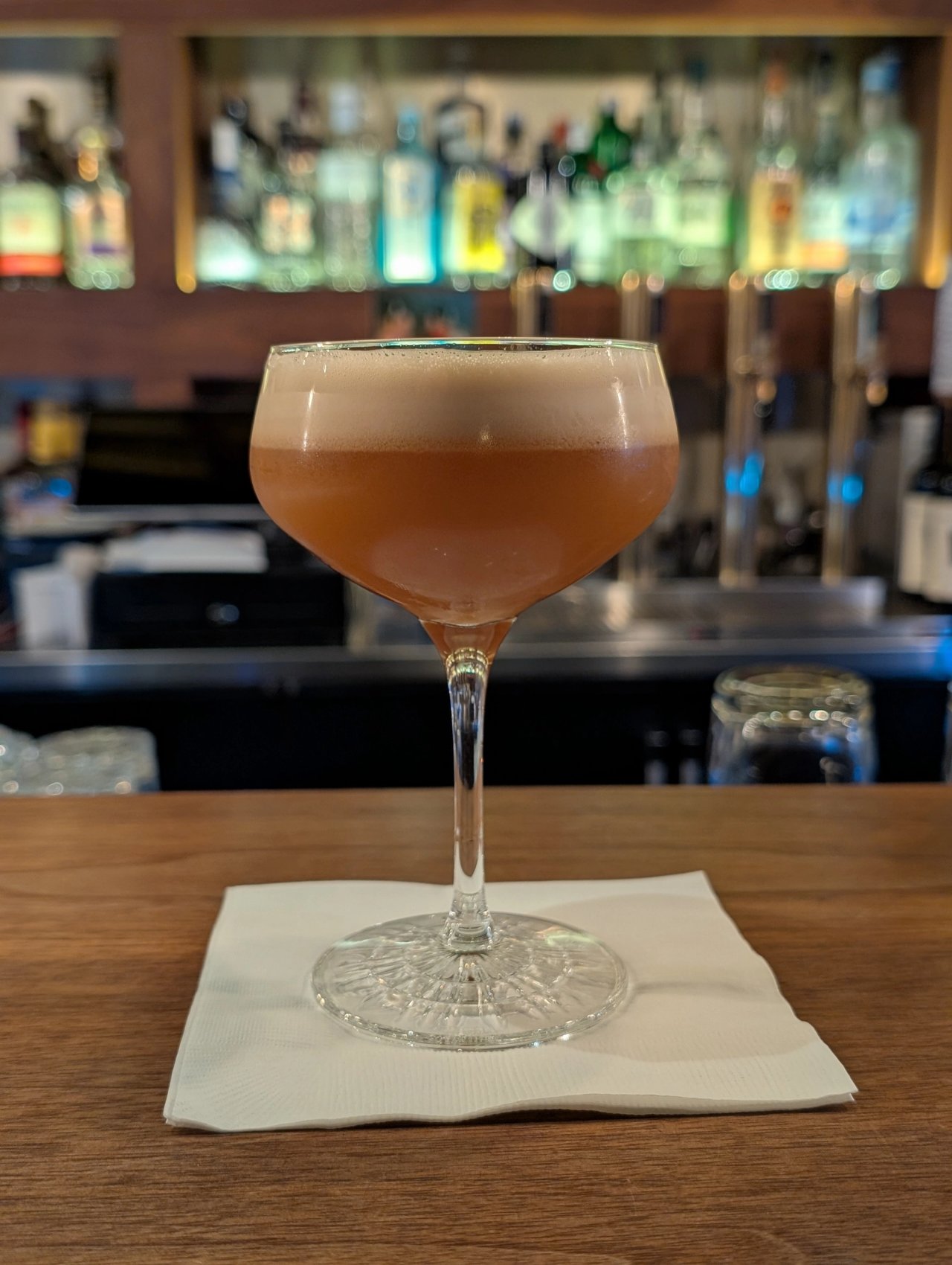 An amber cocktail with a thick foam cap in a stemmed glass at The Narrows, with bottles behind the bar