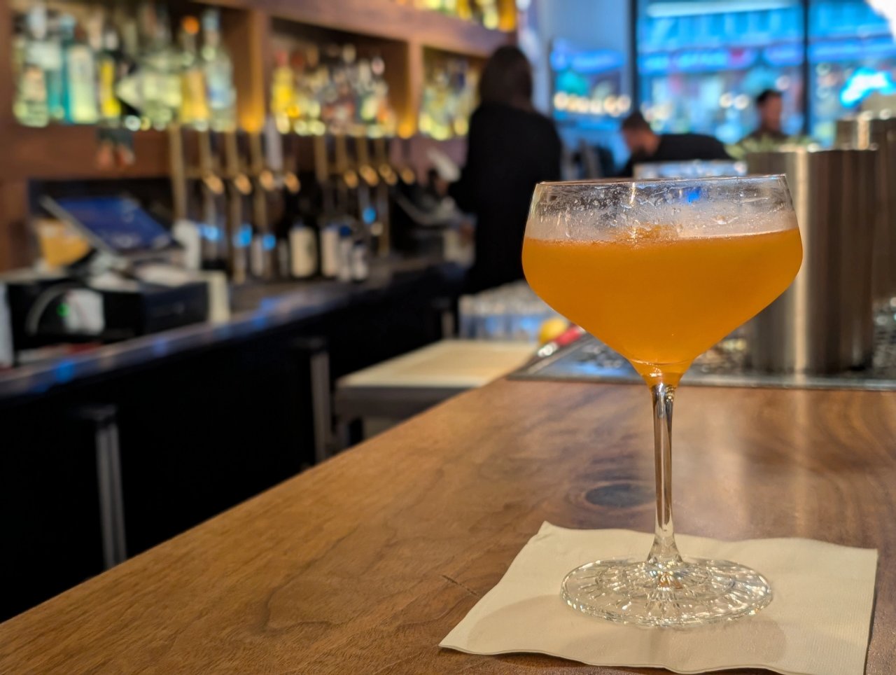 A golden cocktail in a stemmed glass on the wooden bar at The Narrows, with the back bar in view