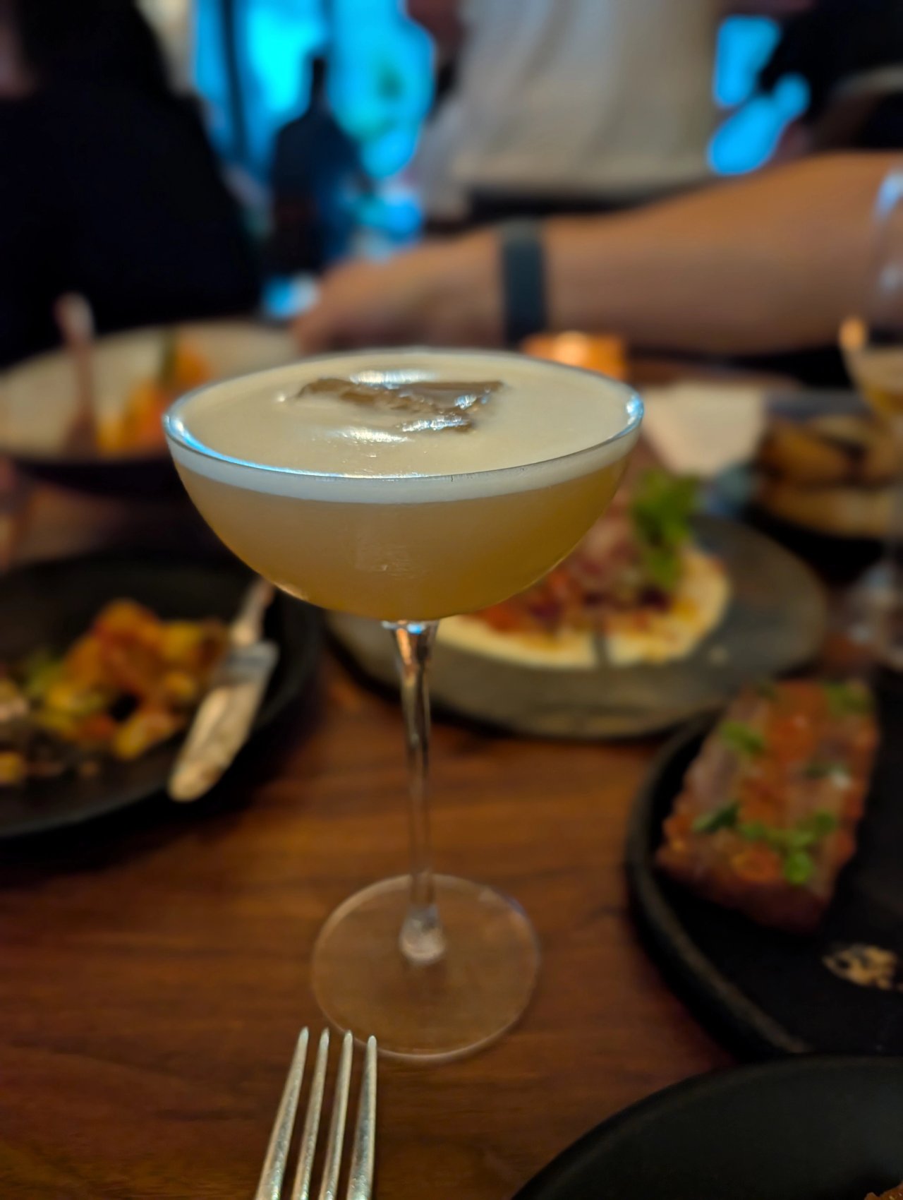 A golden cocktail with a foam cap in a coupe at Porzana, with dishes on the table behind it