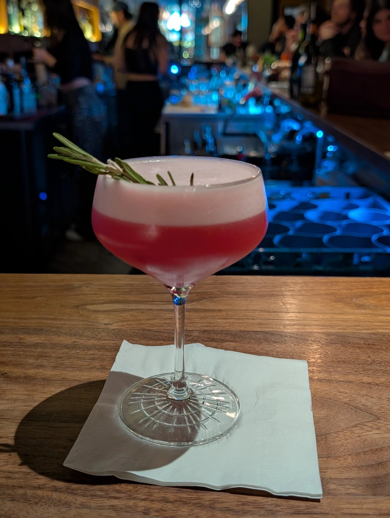 A pink cocktail with a white foam cap and rosemary sprig in a stemmed glass at The Narrows