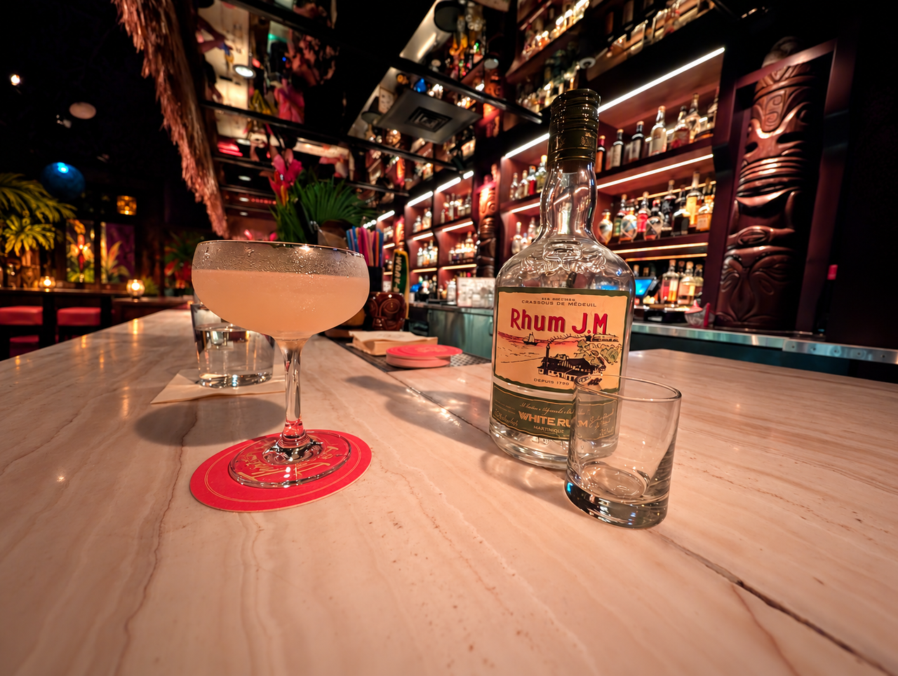 A Hemingway daiquiri and Rhum J.M. bottle on the marble counter, with the illuminated tiki bar behind them