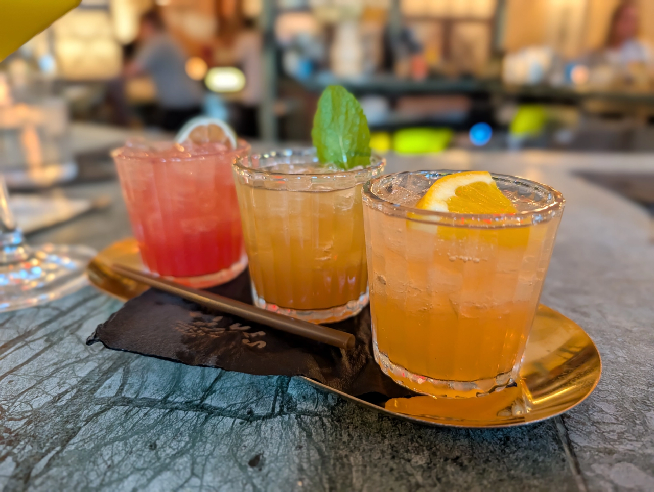 Three small colorful cocktails on a gold tray at Fives, with citrus and mint garnishes.