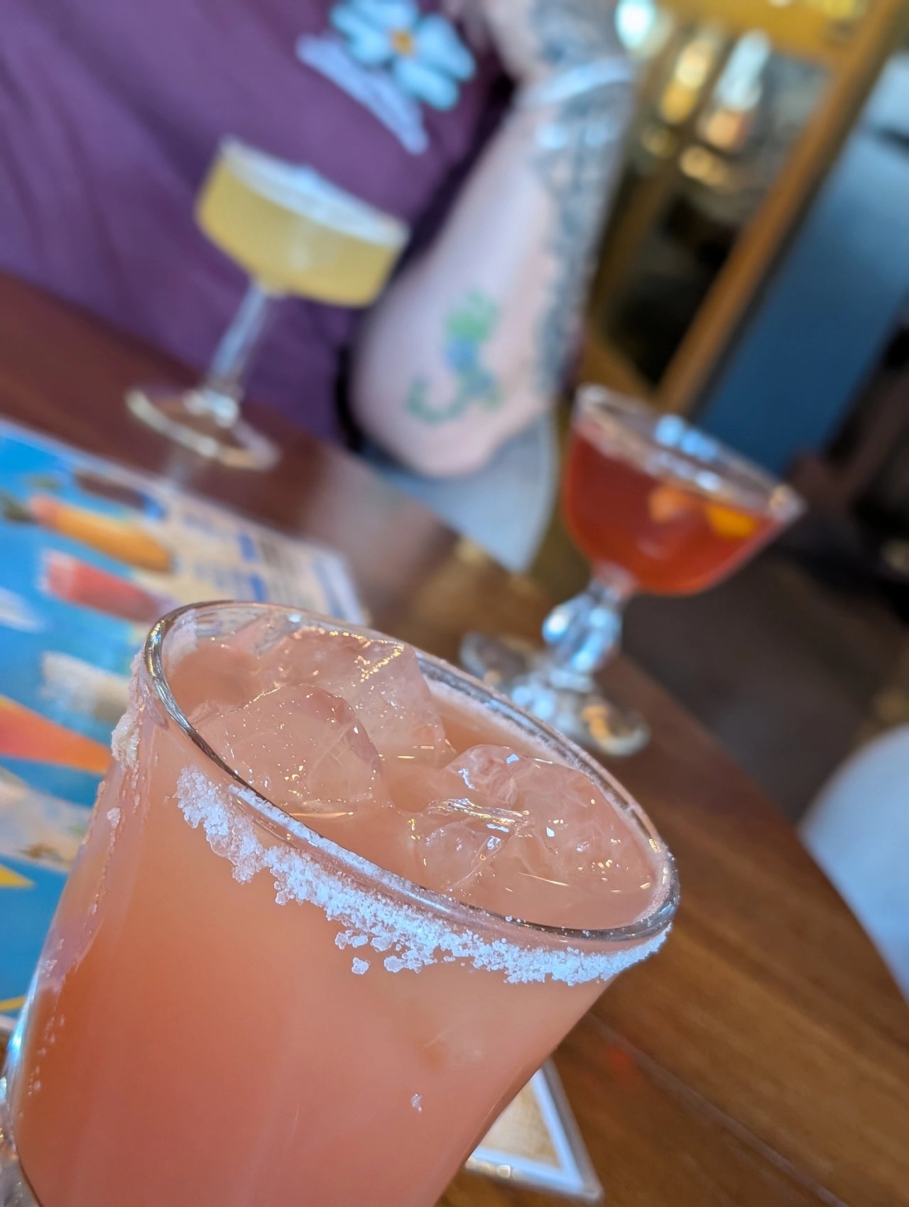 A pink cocktail over ice in a salt-rimmed tall glass at Vikre, with two stemmed cocktails behind it
