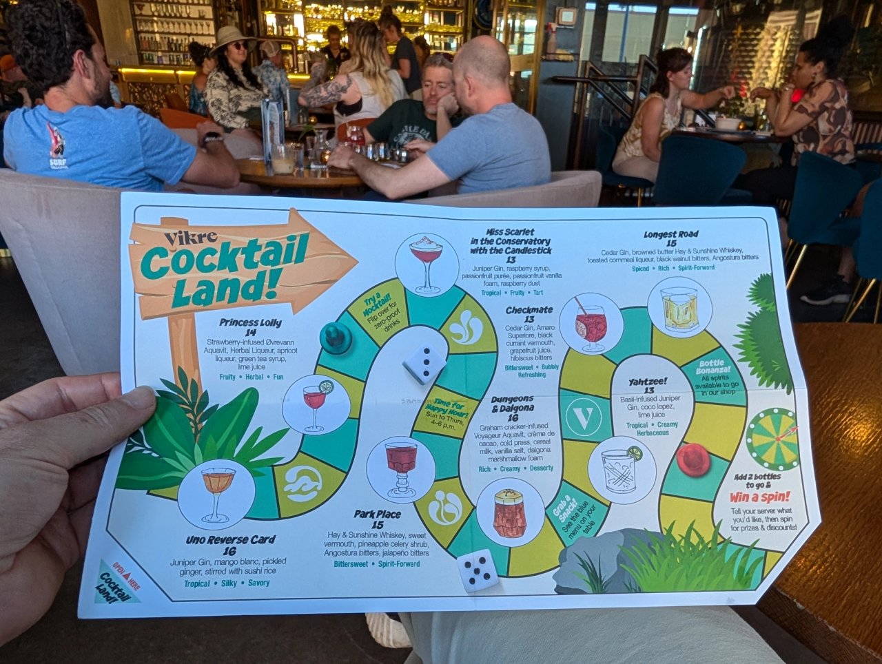 Vikre’s Cocktail Land board-game menu held up in the busy tasting room, with dice and illustrated drinks