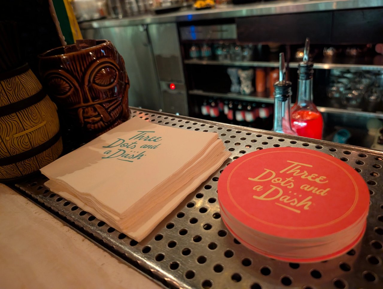Three Dots and a Dash napkins and red coasters beside tiki mugs at the bar