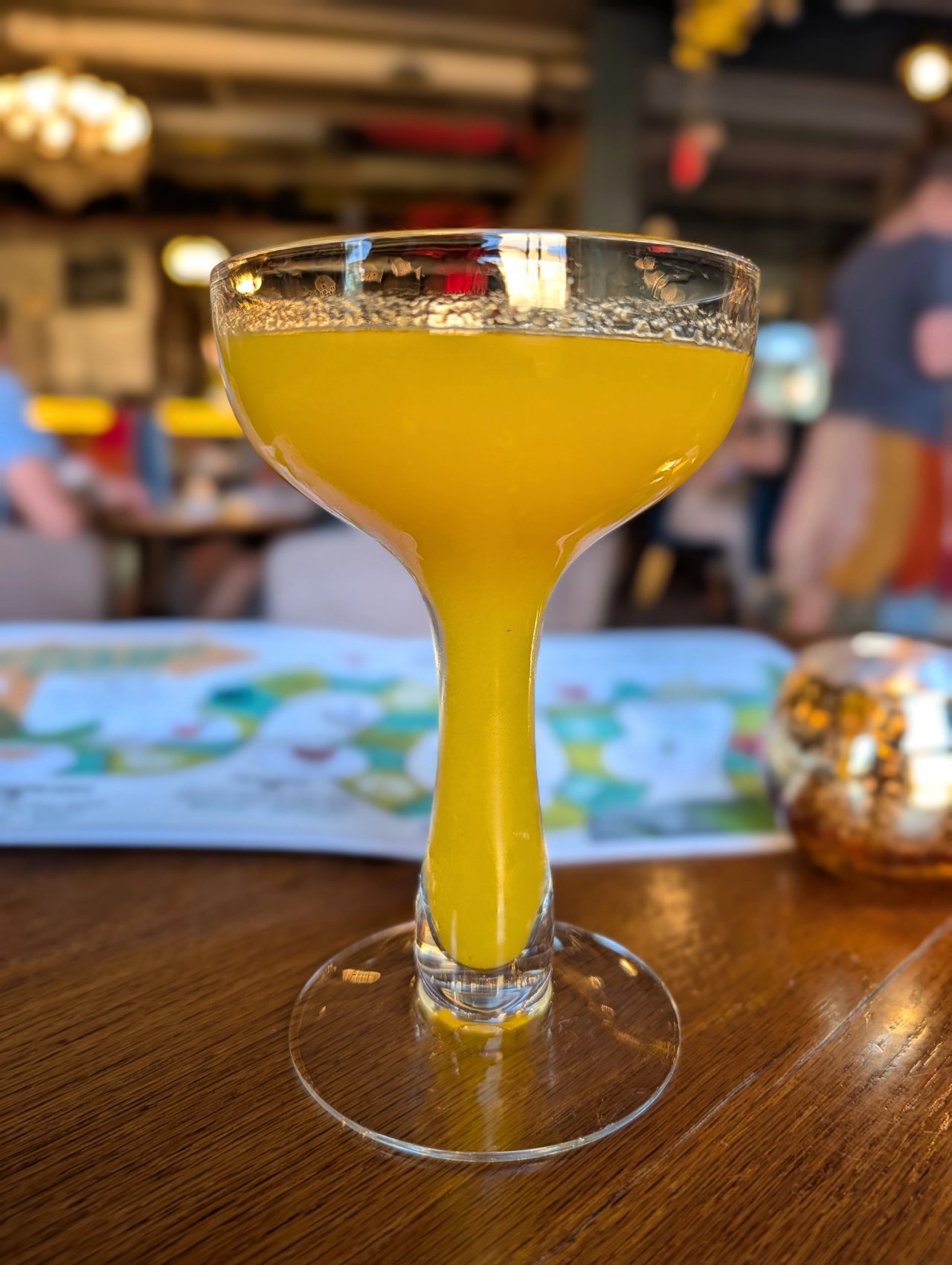 A vivid yellow cocktail in a tall-stemmed glass at Vikre, with a colorful menu behind it