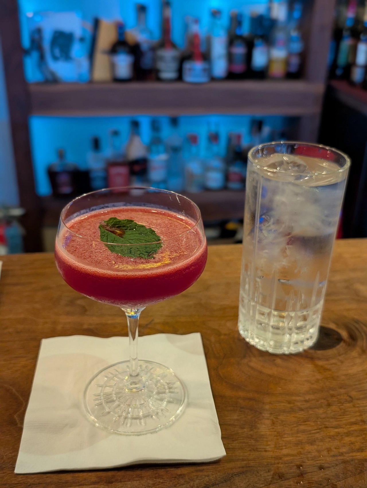 A deep red cocktail with a mint leaf in a stemmed glass beside a tall glass of water at The Narrows