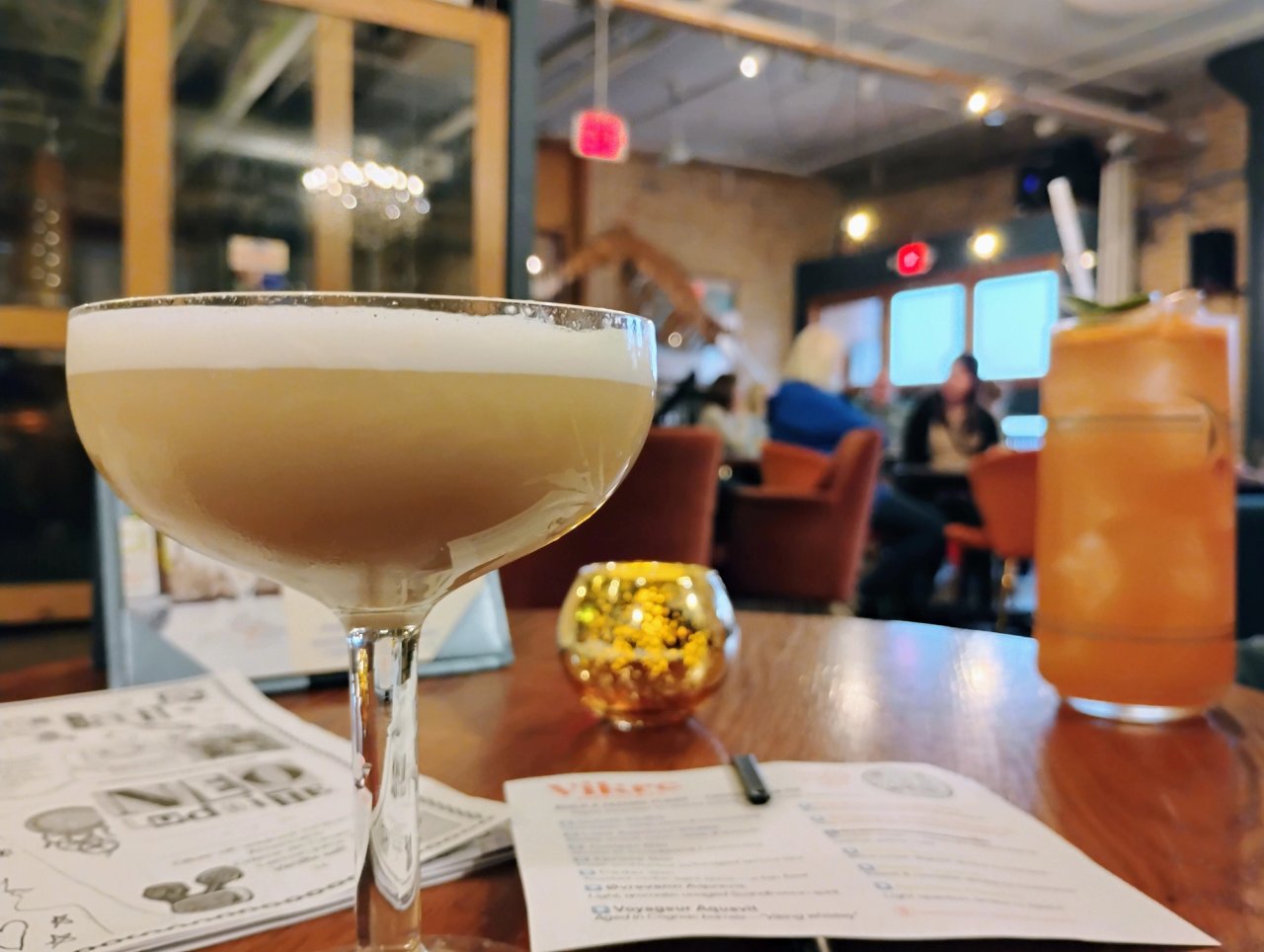 A pale foamy cocktail in a coupe and an orange drink over ice on a table in Vikre’s tasting room