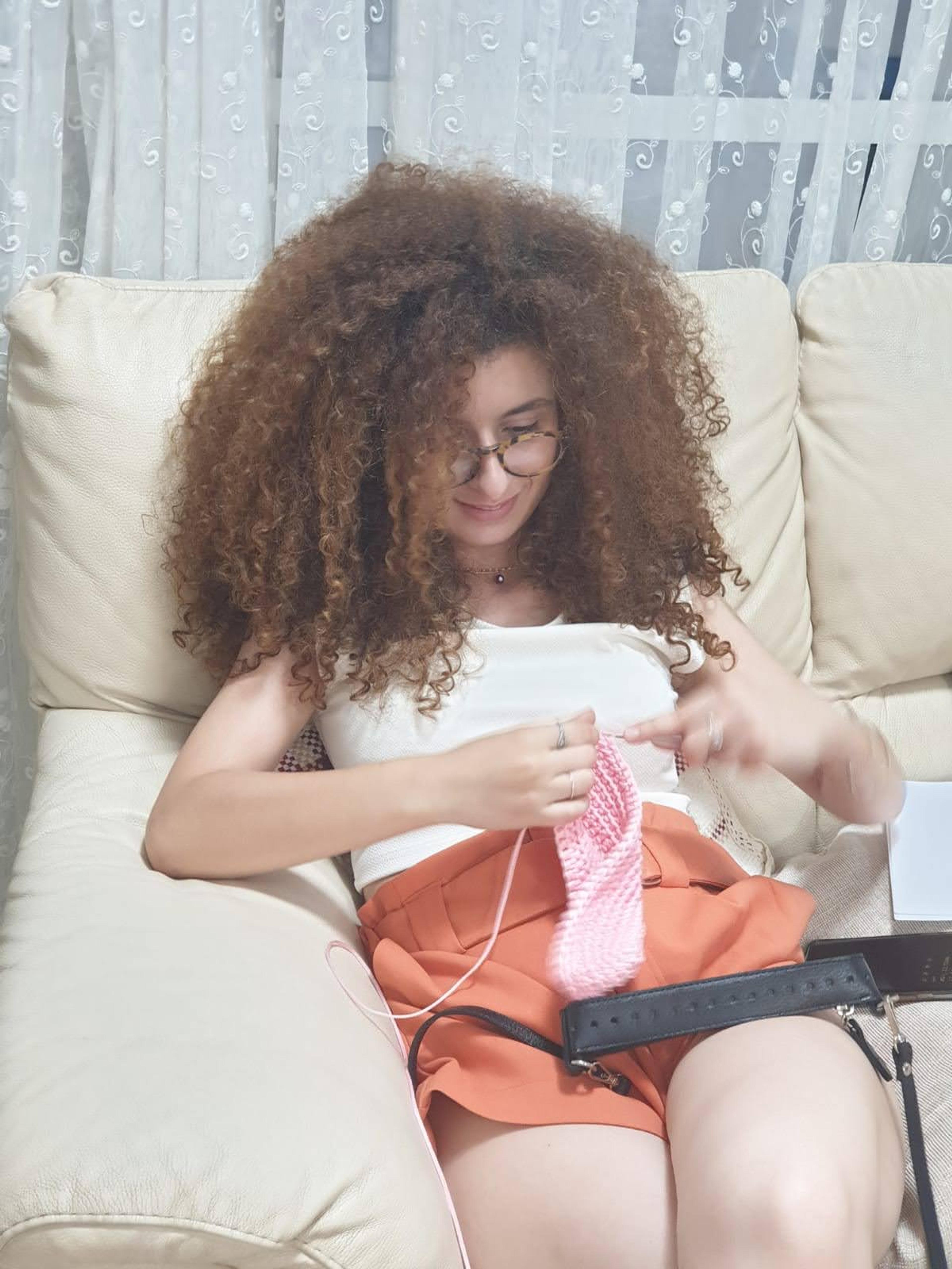 Aluma sitting on a couch and crocheting a crossbody pink bag