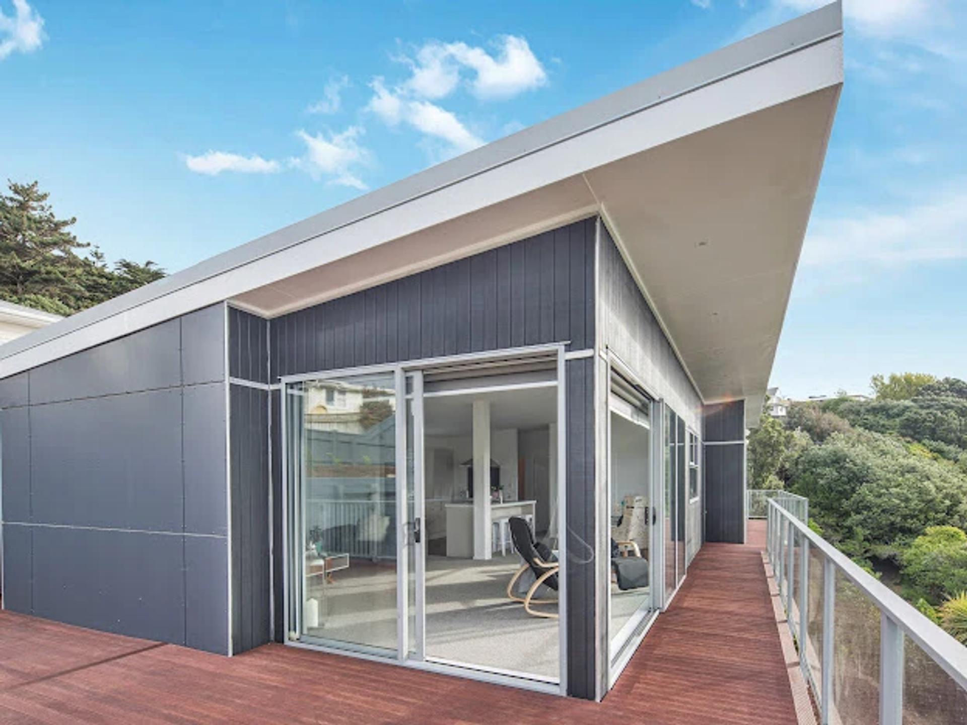 Modern dark gray house with a wooden deck, sliding glass doors, and a view of lush trees.
