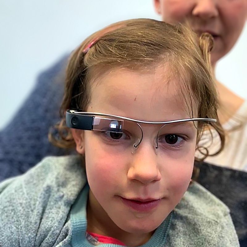 ein kleines Kind mit einer munevo-Brille und einem Elternteil im Hintergrund