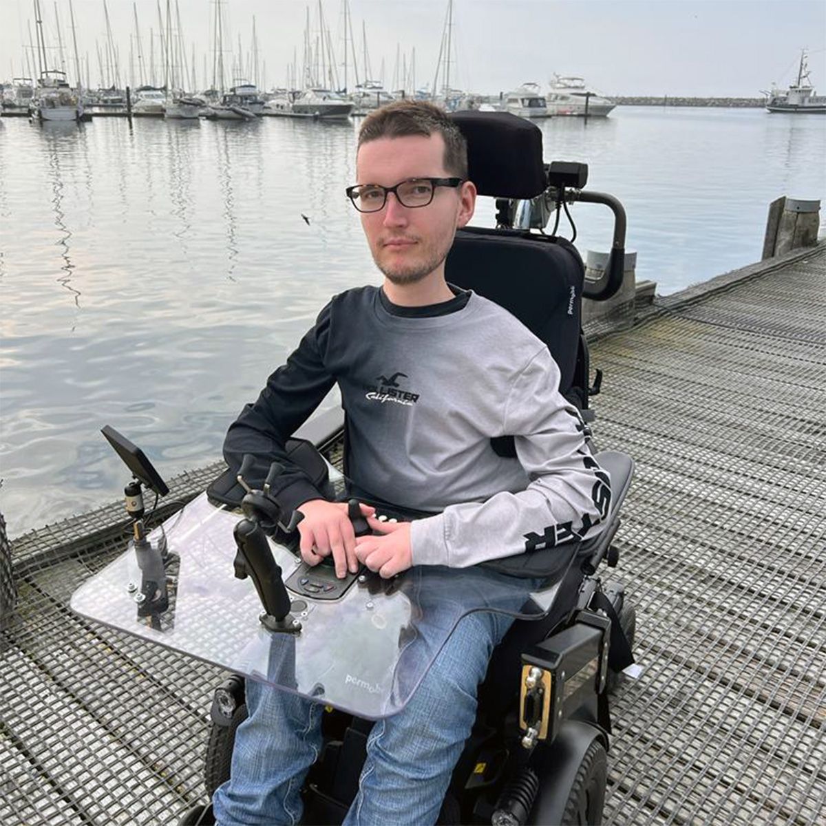 Marcel sitting in his wheelchair on a dock with sailboats in the background