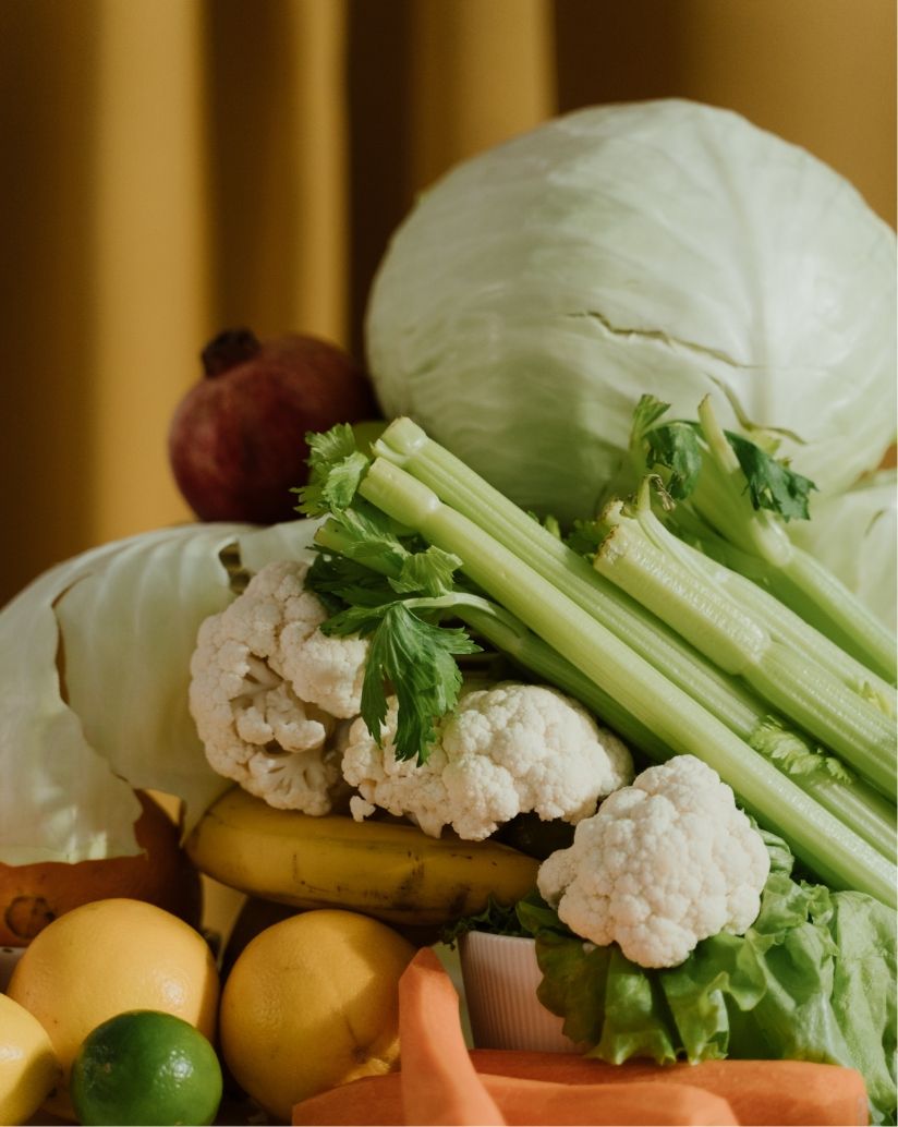 Artful still life photo of groceries and raw produce, including a head of cabbage, celery stalks, heads of cauliflowers, carrots, some grapefruits and a lime