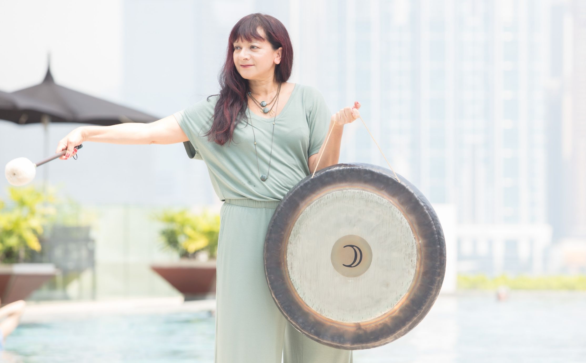 A woman in a green top and pants holding a gong in one hand and a mallet with a stretched out hand in the other
