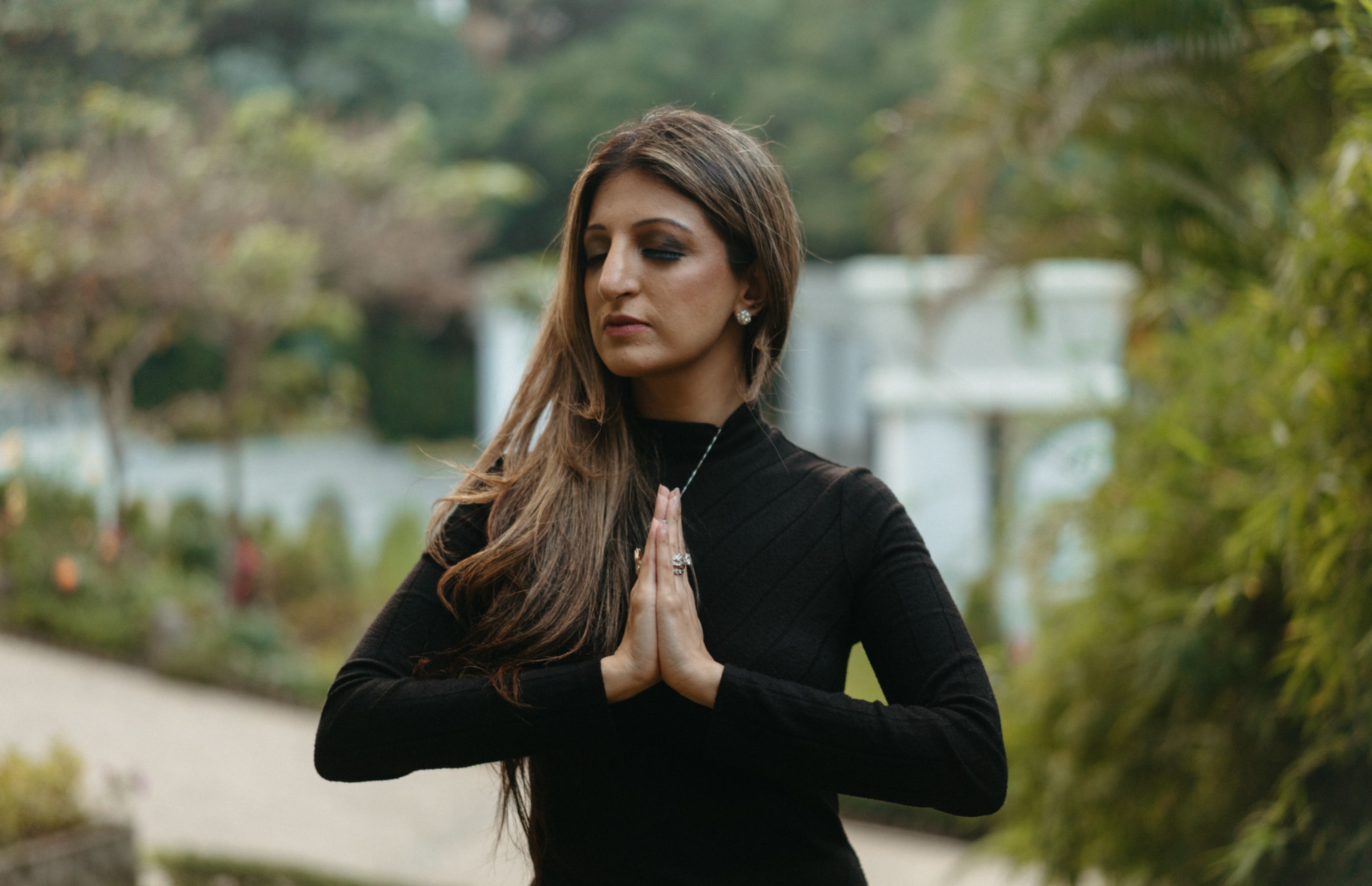 Woman with long hair in black top with hands together in prayer pose in front of her chest, looking peaceful with eyes closed