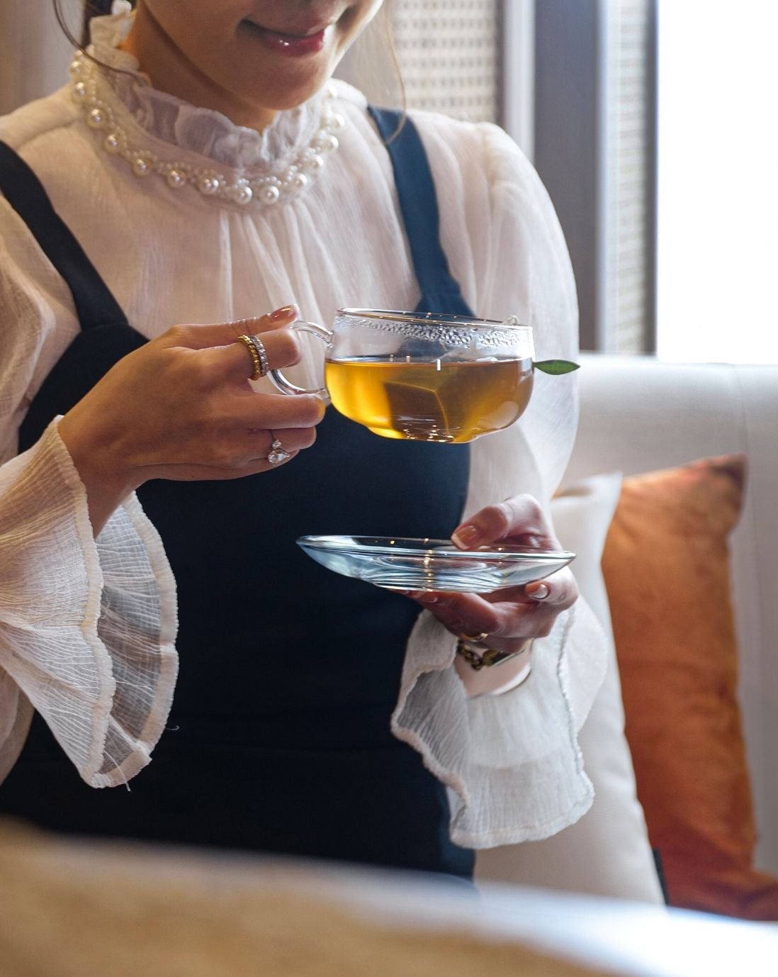 A woman in white blouse and black dress sitting on couch and drinking tea from transparent tea cup in one hand and saucer in the other
