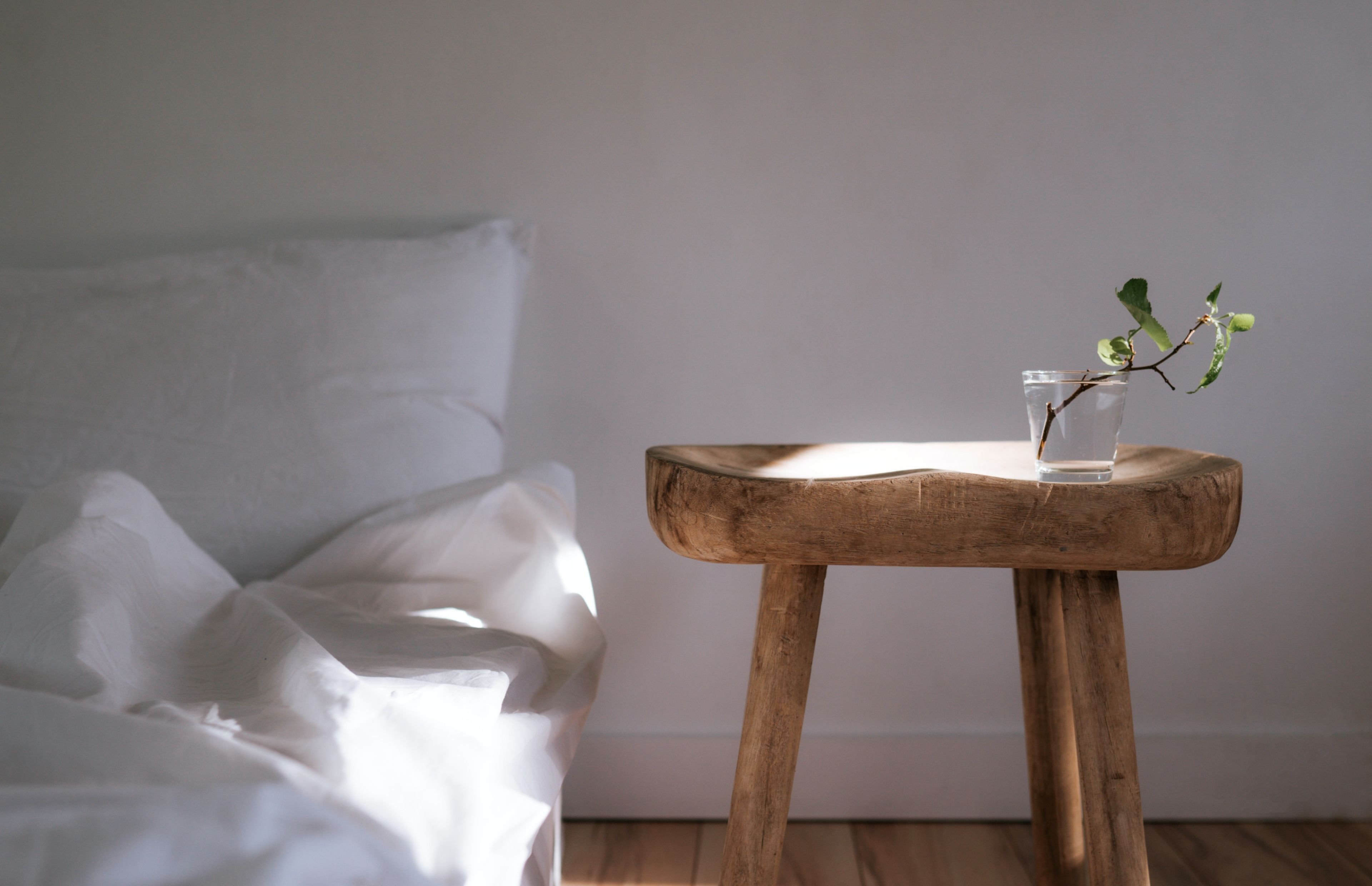 A glass cup filled with later with a branch on a wooden stool. by a white bed