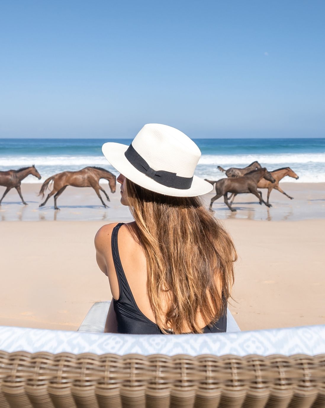 Woman in hat and bathing suite sitting by the ocean with horses running along the shore