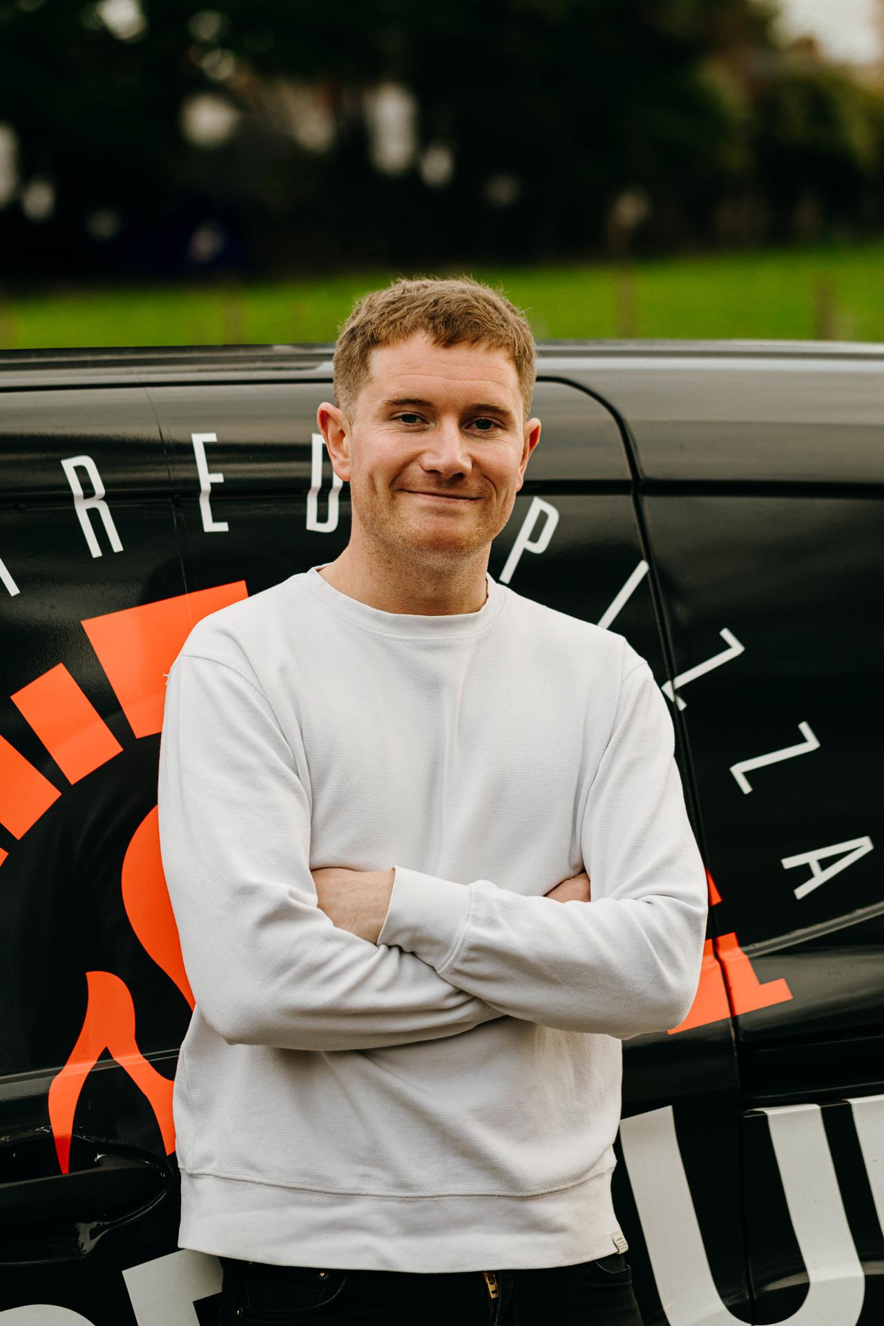 A smiling Fired Up Pizza team member standing in front of a delivery van