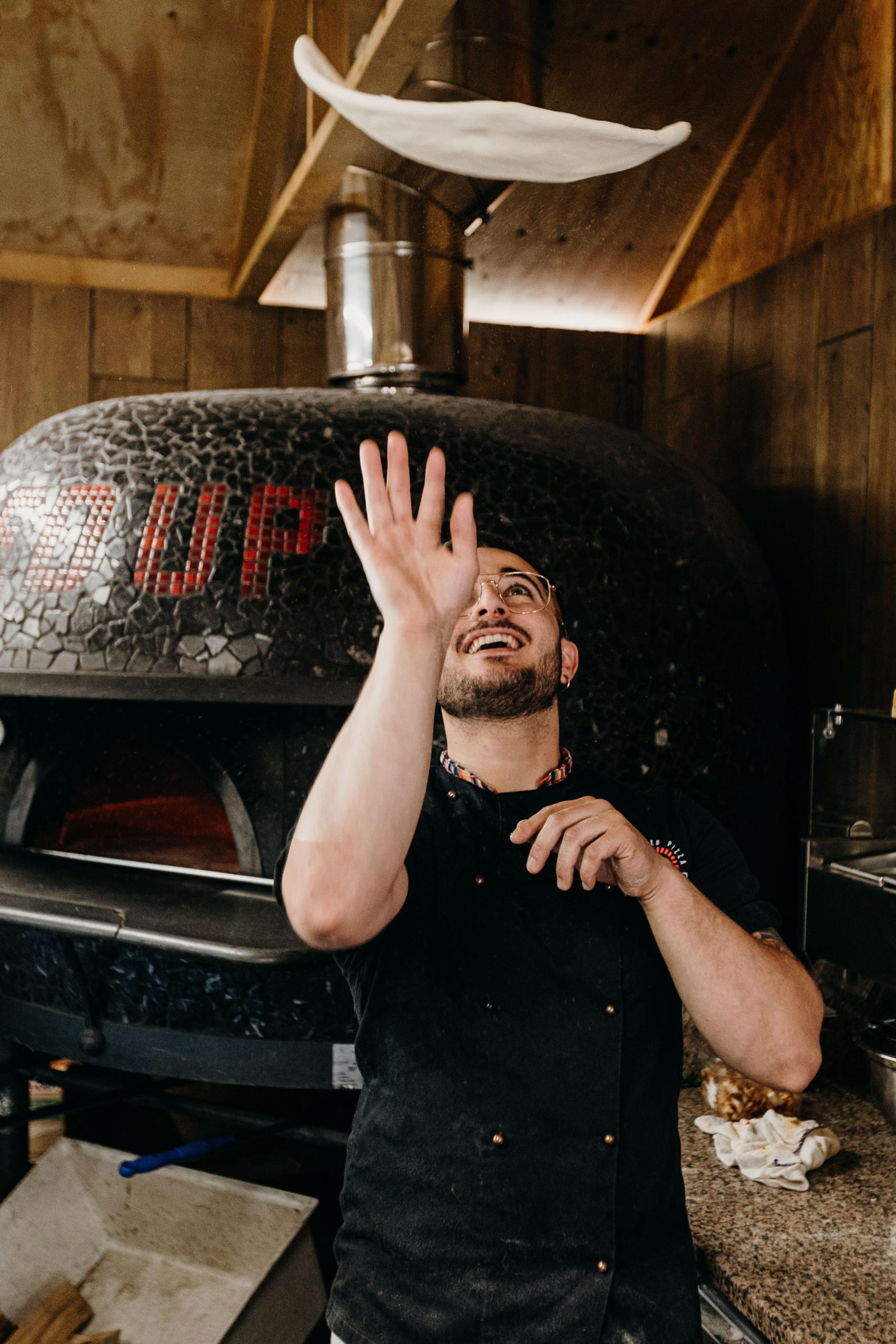 A Fired Up pizza maker tossing pizza dough in front of a wood-fired oven