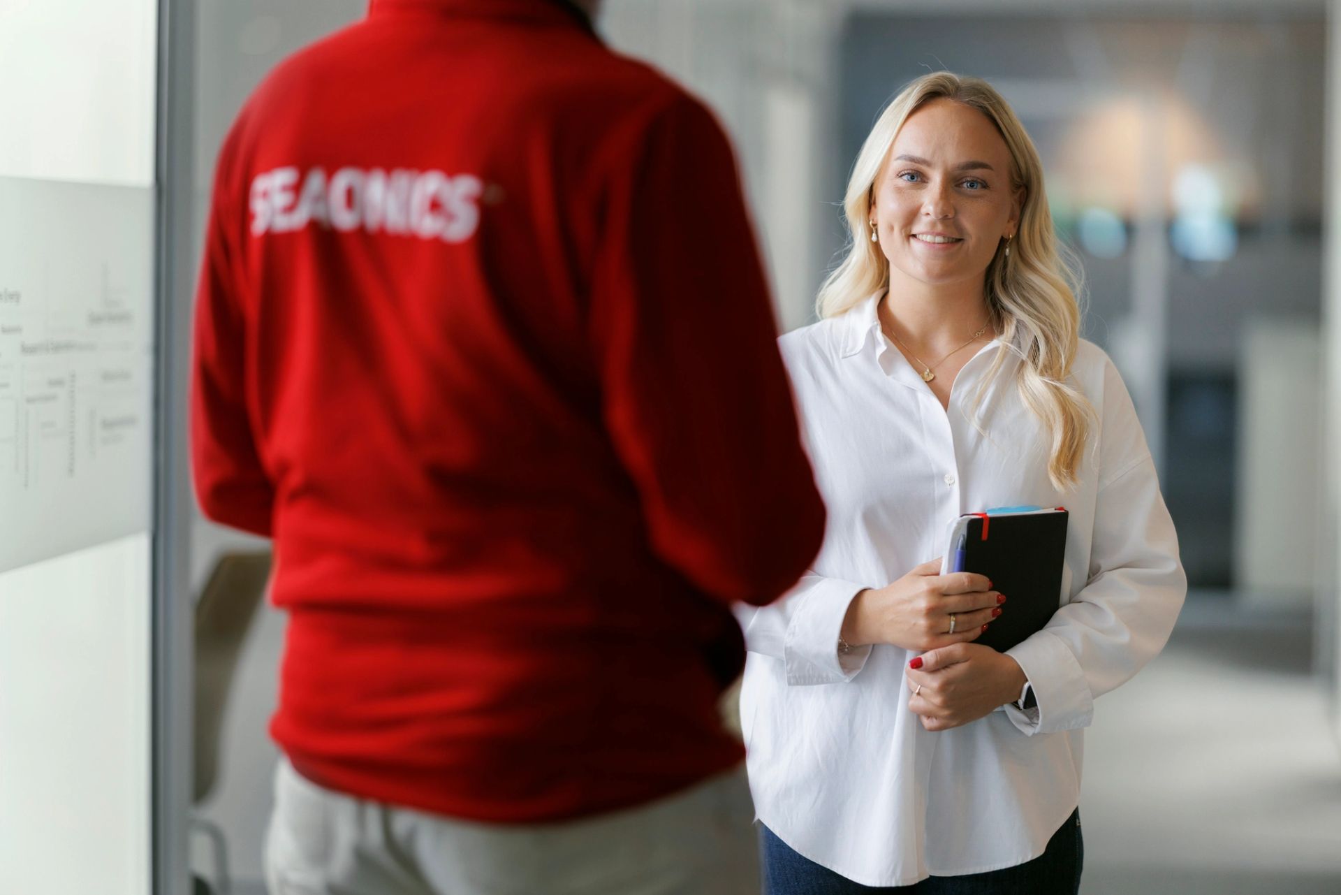 Smiling woman in white blouse holding documents, talking with a colleague wearing a red Seaonics jacket in a modern office environment.