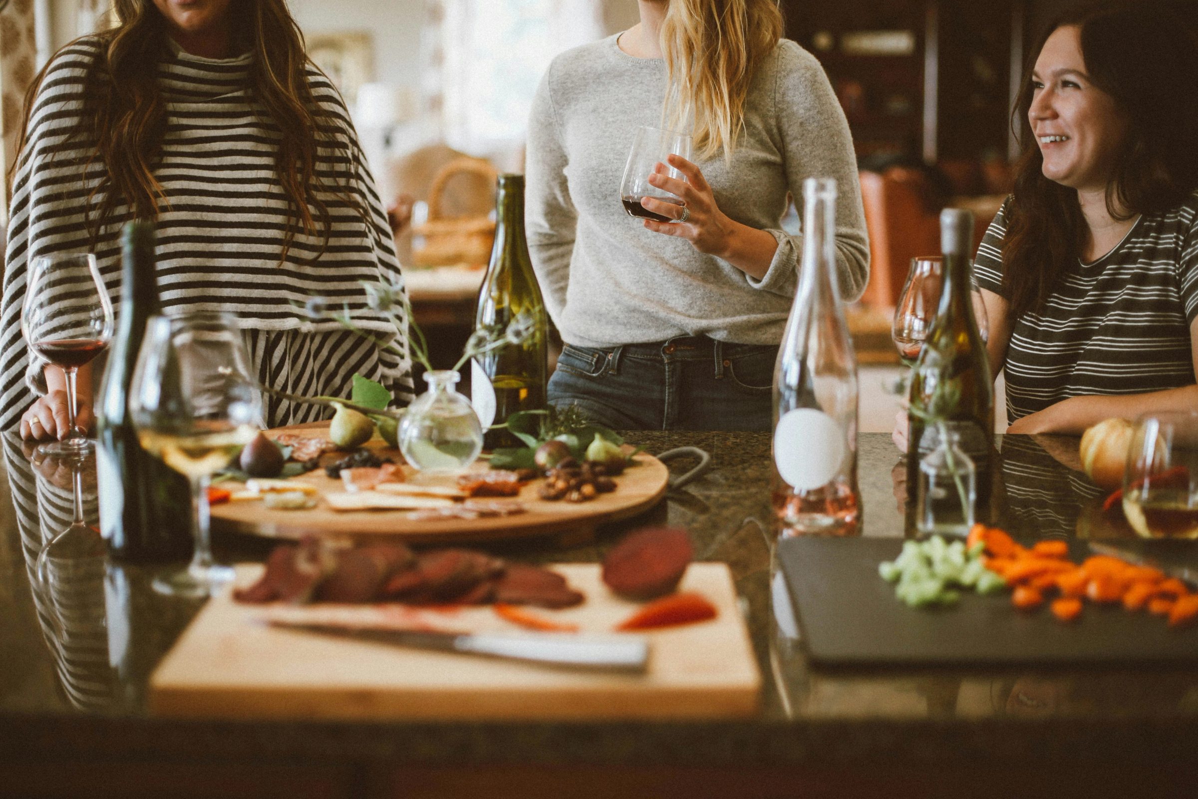 Three people stand at a counter with food, drinks and glasses. Cutting boards display chopped vegetables and ingredients.