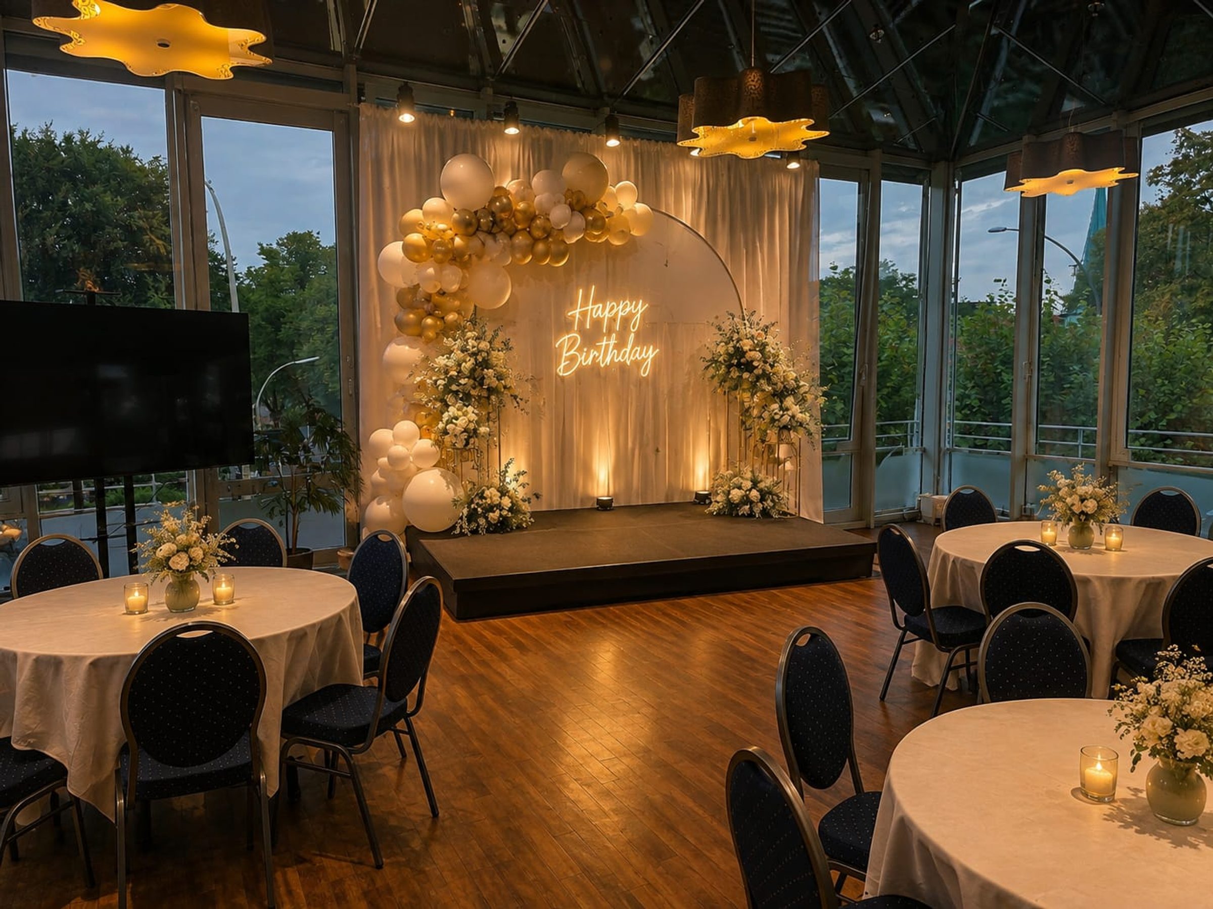 Veranstaltungsraum mit Holzparkett, runden Tischen, weißen Tischdecken und schwarzen Stühlen. Bühne mit Neonschrift "Happy Birthday", Ballondekoration und Blume