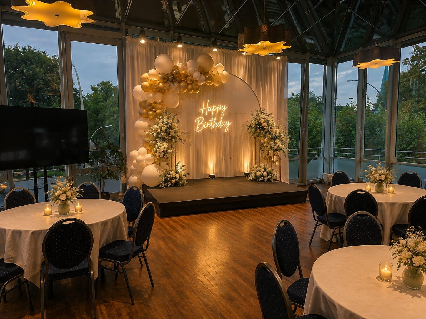 Veranstaltungsraum mit Holzparkett, runden Tischen, weißen Tischdecken und schwarzen Stühlen. Bühne mit Neonschrift "Happy Birthday", Ballondekoration und Blume