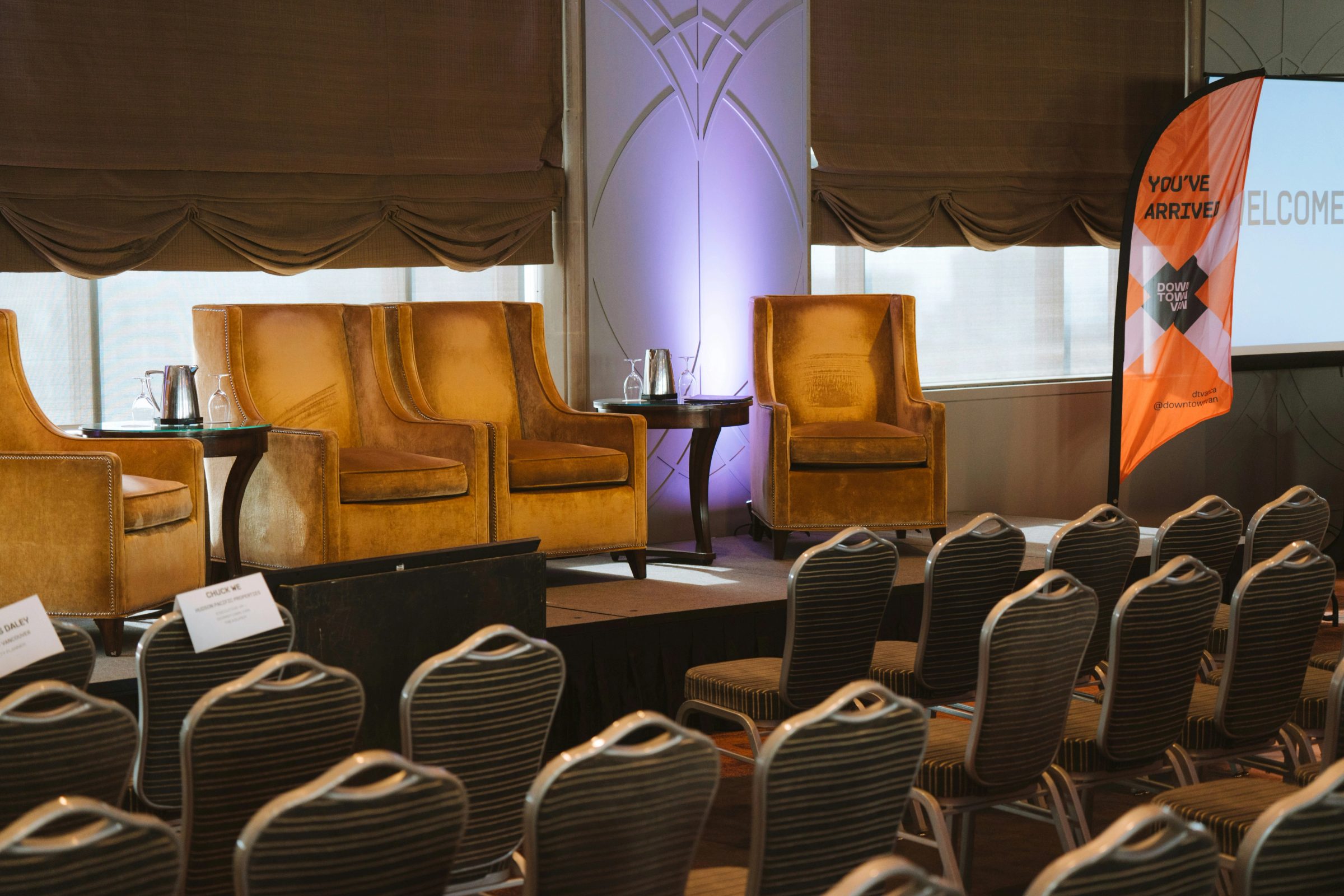 Event space with rows of chairs in foreground, orange leather armchairs on stage, and purple-lit pillar in center.