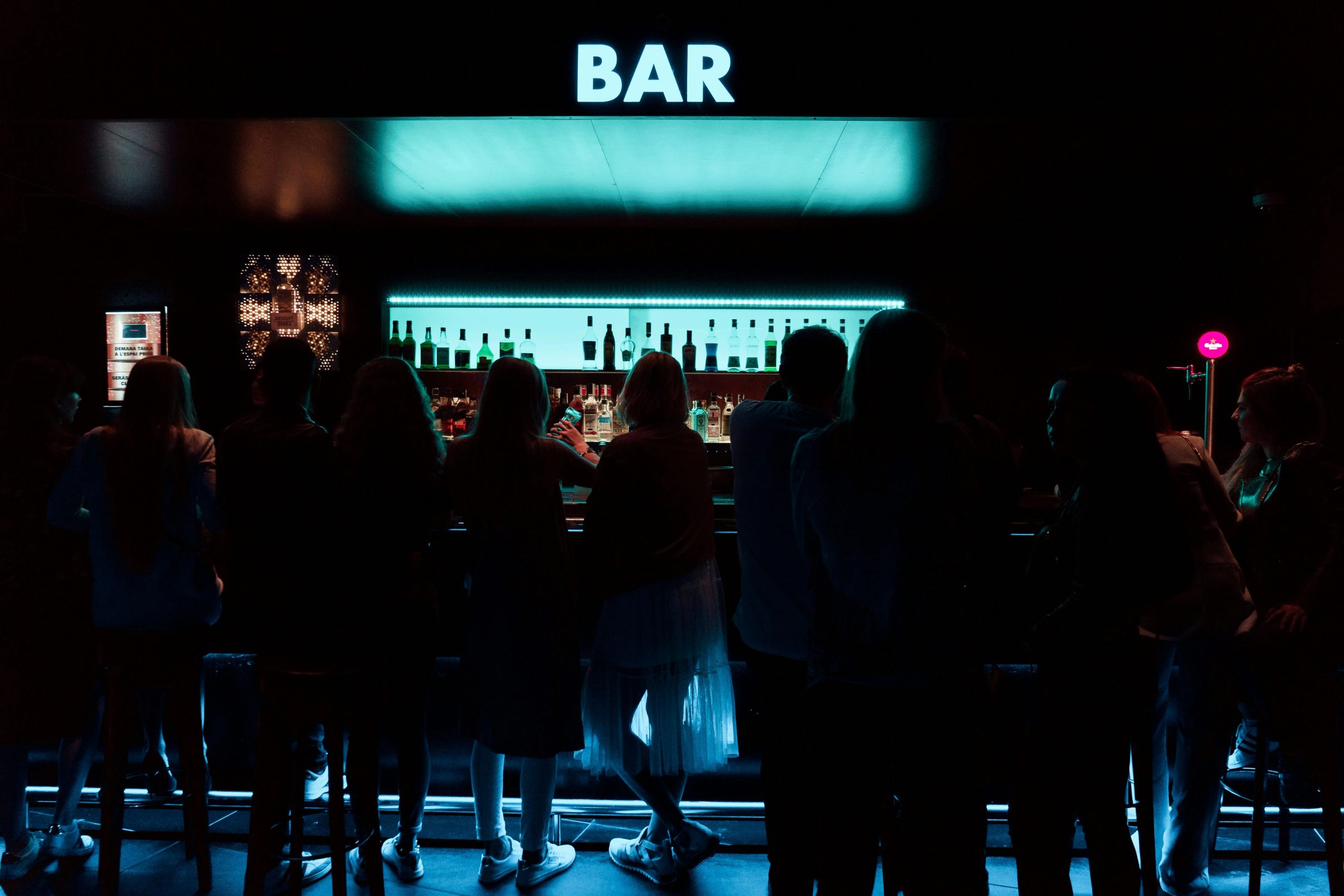 Modern bar with turquoise neon lighting, bottle shelves, and guests at the counter, dimly lit with atmospheric ambiance.