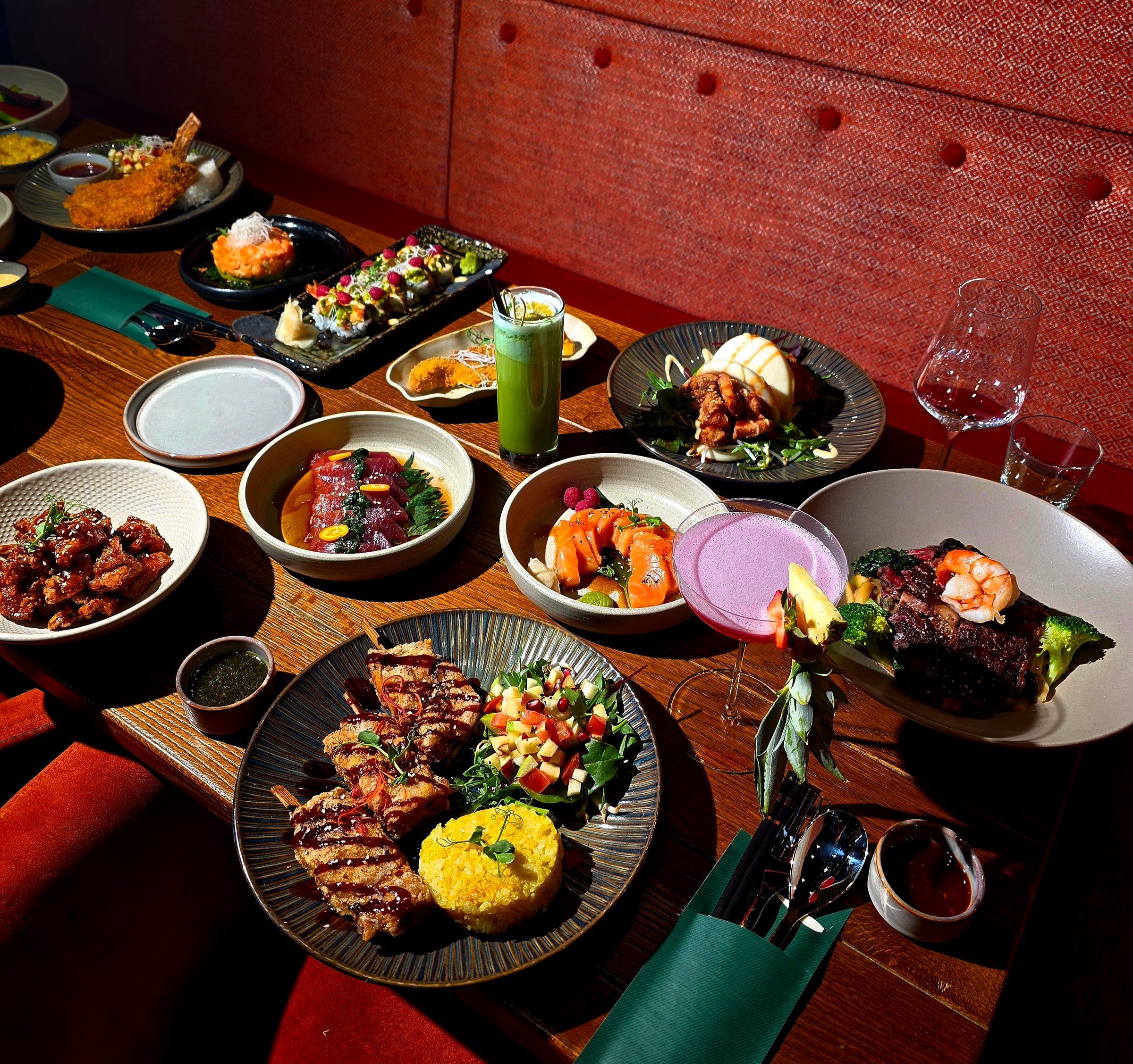 Wooden table with assorted Asian dishes in bowls and plates, red upholstered seating in background, glasses with beverages
