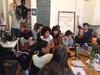 A workshop situation is shown. Fifteen young adults, Black and white, sit on the floor and on chairs in a room. On the walls hang a written board and a handwritten poster. It reads: We are born free. Empowerment. Radio and Coffee. Other smaller pieces of paper can be seen on the wall. Two people are raising their hands. 